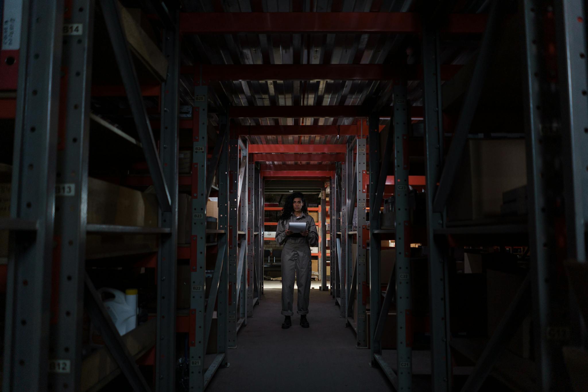 A woman stands in a narrow warehouse corridor, inspecting inventory under dim lighting.